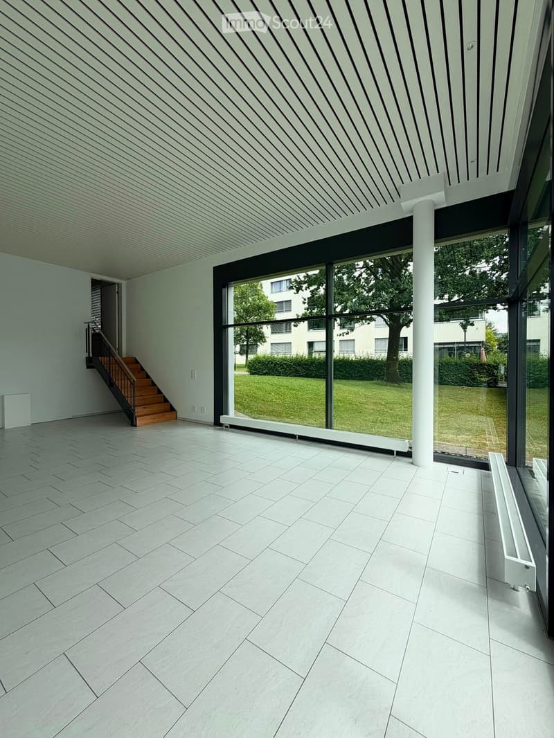Bright, minimalist room with large glass windows, white tiled floor, and a wooden staircase leading upwards.