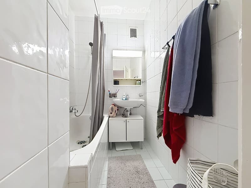 A small, modern bathroom with white tiled walls and floor, featuring a bathtub, sink with a mirror, a small cabinet, and towels hanging on a rack.
