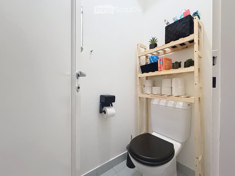 A compact bathroom with a toilet, a wooden shelf unit holding toilet paper, small plants, and other items, and a toilet paper holder on the wall.