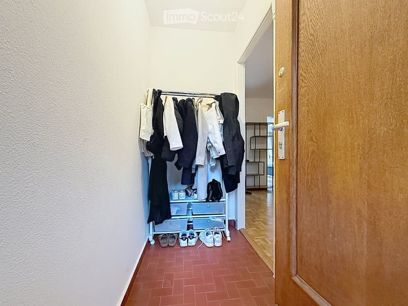 A narrow hallway with a coat rack holding jackets and coats, a shoe rack with shoes, and a small shelf unit visible in the background.