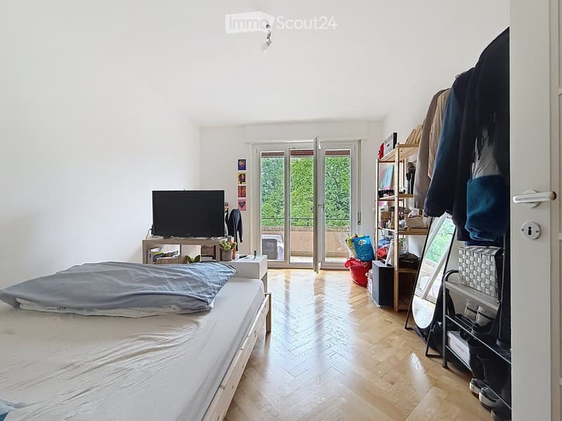 Bright bedroom with wooden flooring, a bed with pillows, a TV on a stand, a shelf with books and items, and a mirror leaning against the wall. The room has large windows and a sliding glass door leading to a balcony.