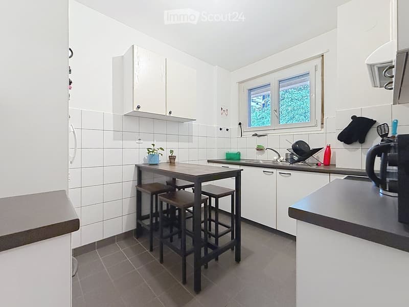 Modern kitchen with white cabinets, tiled walls, and a central island table with four stools. The kitchen features a sink, oven, and various small appliances and utensils.