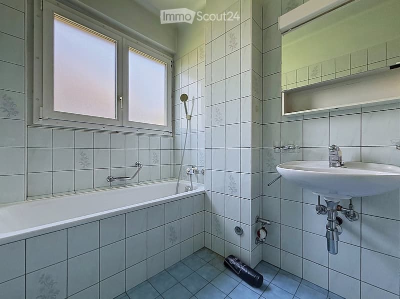 Bathroom with white tiled walls and floor, featuring a bathtub, a sink, and a showerhead. There is a window above the bathtub and a small shelf above the sink.
