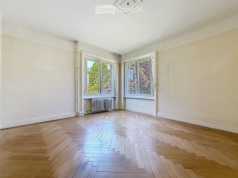 Empty room with wooden herringbone flooring, white walls, large windows, and a radiator under one window.