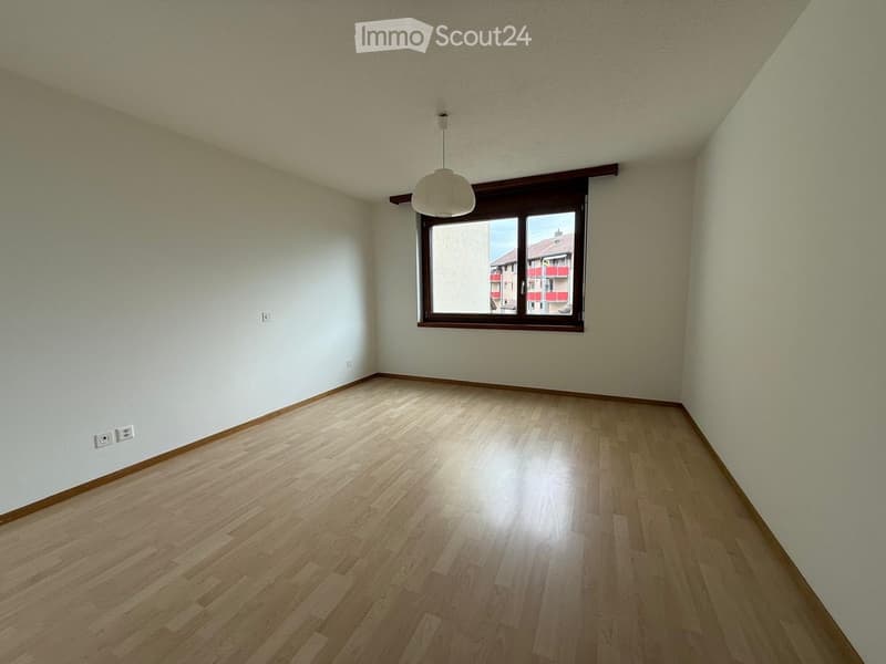 Empty room with light wooden flooring, white walls, and a single window with a view of neighboring buildings.