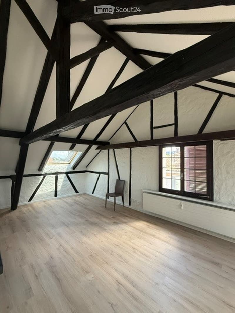 Empty attic room with wooden beams, exposed wooden ceiling, wooden floor, single window, and a single chair.