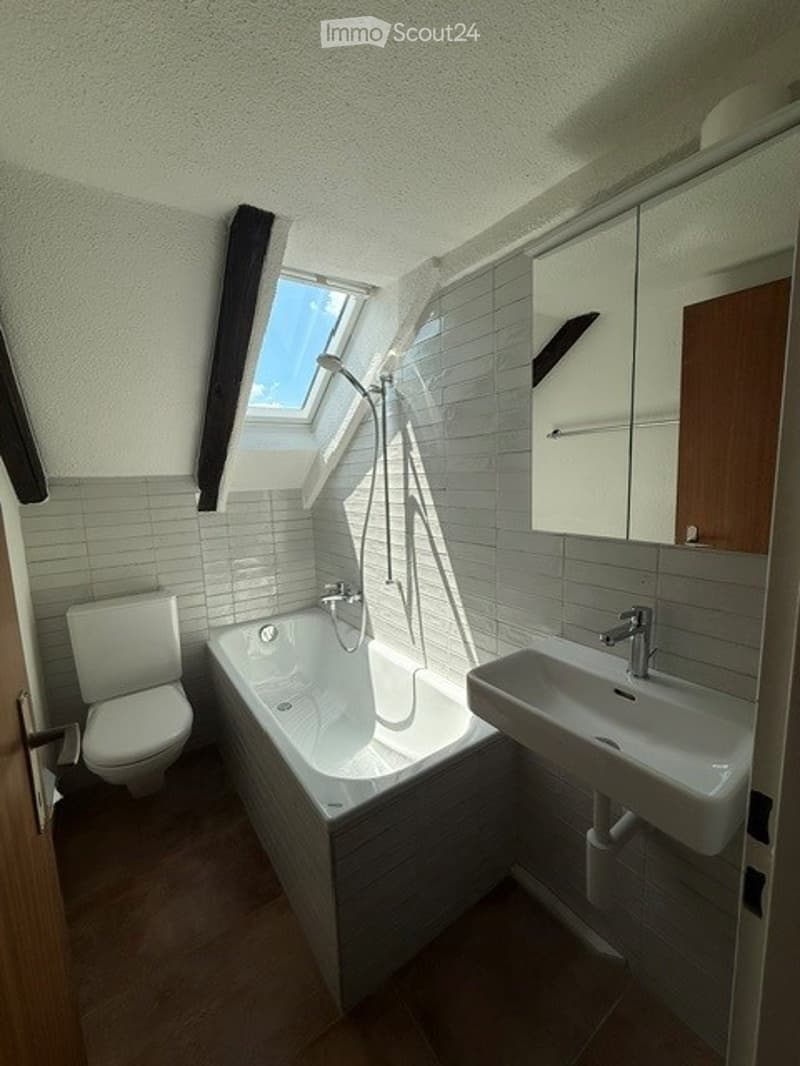 Modern bathroom with a bathtub, toilet, sink, mirror, and a showerhead. The room features white tiles, a skylight window, and a wooden floor.