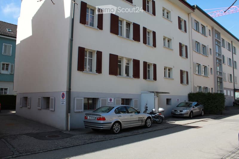 White building with multiple windows, two cars parked, two scooters parked, metal grate on sidewalk