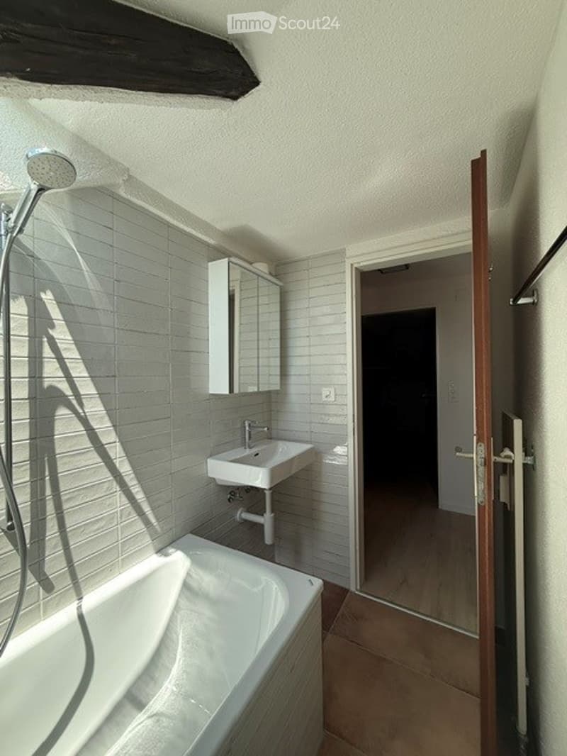 Modern bathroom with a white bathtub, a white sink, a mirror cabinet, and a showerhead. The walls are tiled in light grey, and the floor is tiled in a warm brown color. An open door leads to another room.