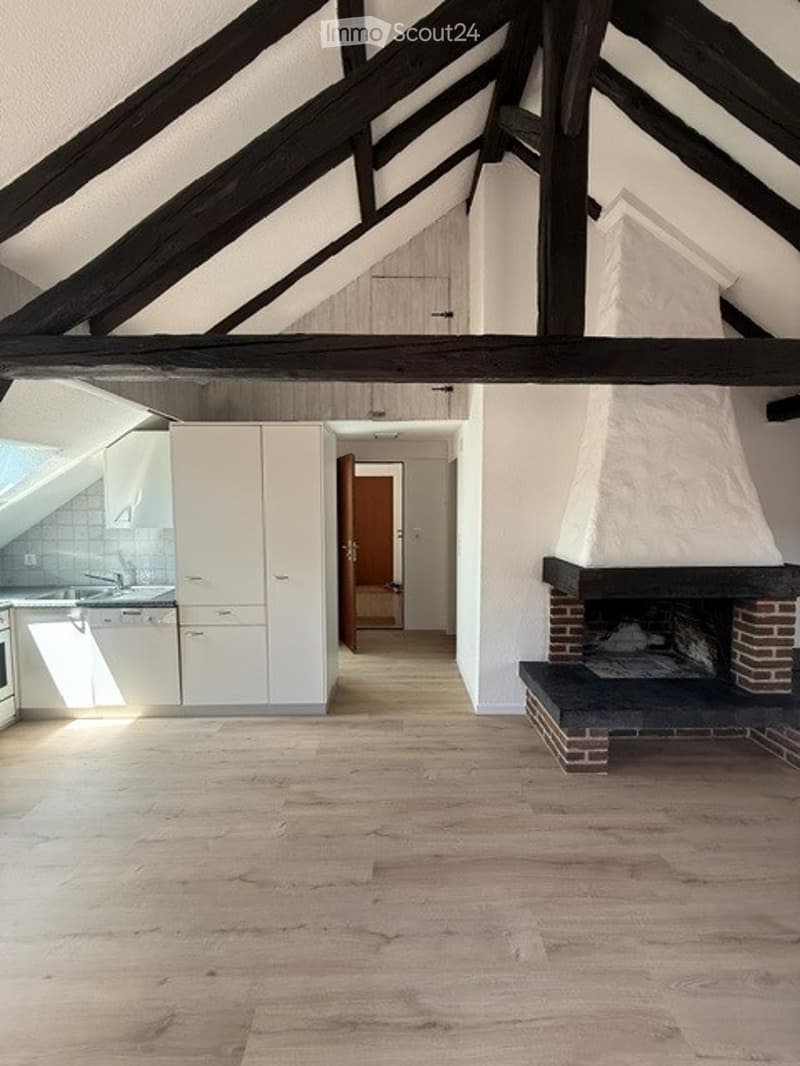 Bright, open-plan attic space with exposed wooden beams, modern white kitchen units, a brick fireplace, and wooden flooring.