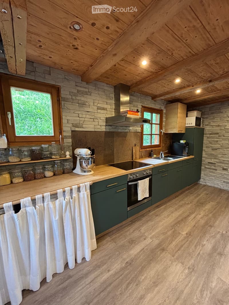 Modern kitchen with wooden ceiling beams, stone backsplash, green cabinets, and a central island with a mixer and jars. Features an oven, sink, and microwave.