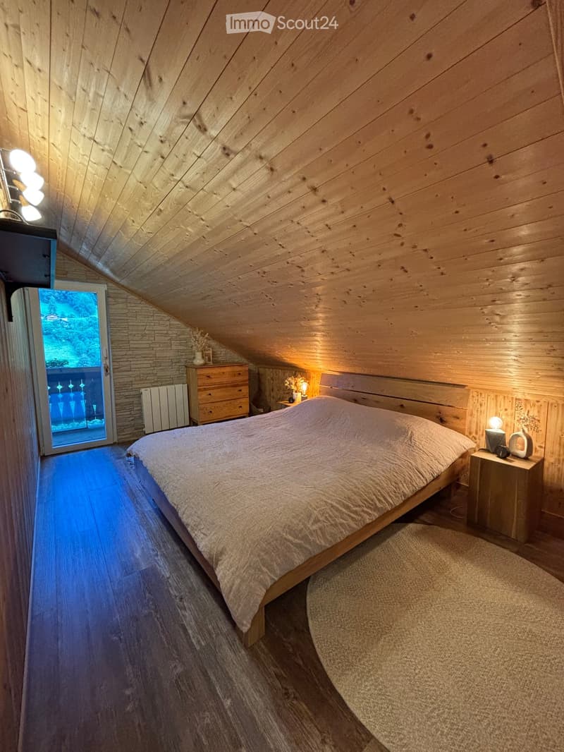 Cozy attic bedroom with wooden walls and slanted ceiling, featuring a large bed, nightstands, lamps, and a view through a glass door.