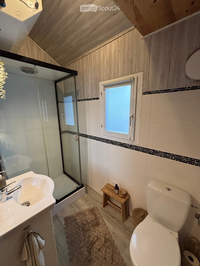 Modern bathroom with a sink, toilet, shower, small wooden stool, and a window letting in natural light.