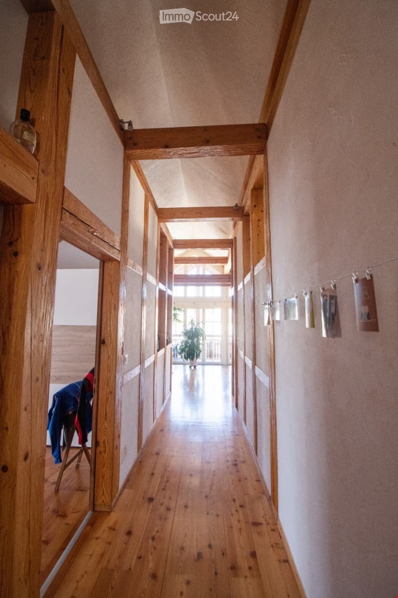 A long wooden hallway with exposed wooden beams, light-colored walls, and a wooden floor. Natural light streams in from the end of the corridor, and a small plant is visible in the distance.