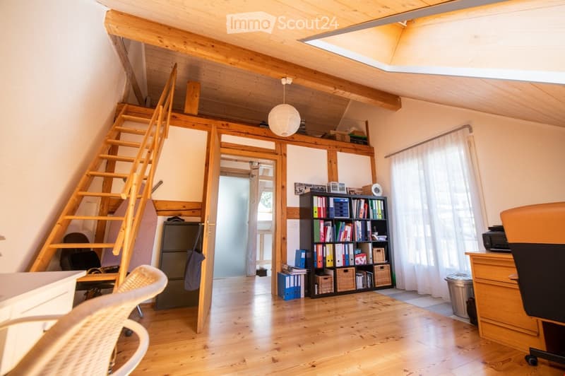 Bright attic room with wooden floors, slanted wooden ceiling, large window with white curtains, and a wooden staircase leading to a loft area. Features a desk with a chair, a bookcase filled with books and binders, a small refrigerator, and a white hangin