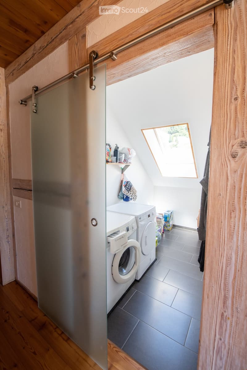 A small, modern bathroom with a washing machine, a shelf with cups and bottles, and a sliding glass door leading to another room.