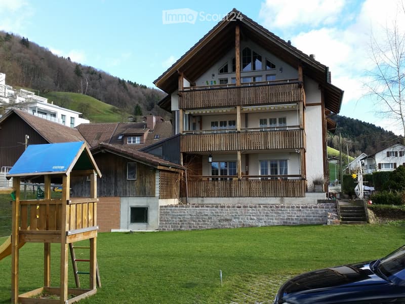 A two-story house with a wooden balcony, surrounded by a green lawn and a wooden play structure. The house features a stone base and wooden upper levels, with multiple windows and a sloped roof.