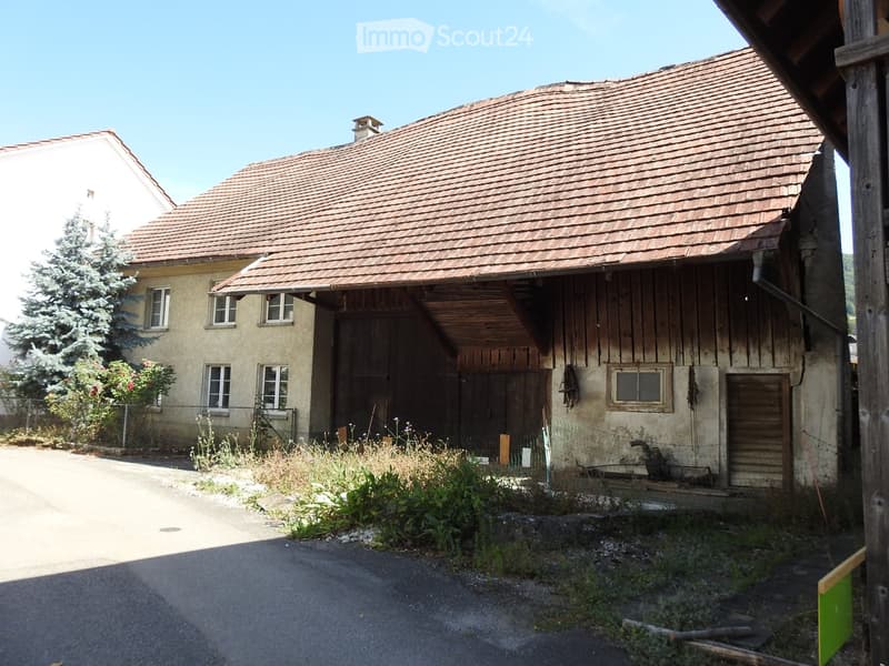 Two-story building with a tiled roof, wooden garage, and weathered exterior walls. The building has multiple windows and a small chimney on the roof.