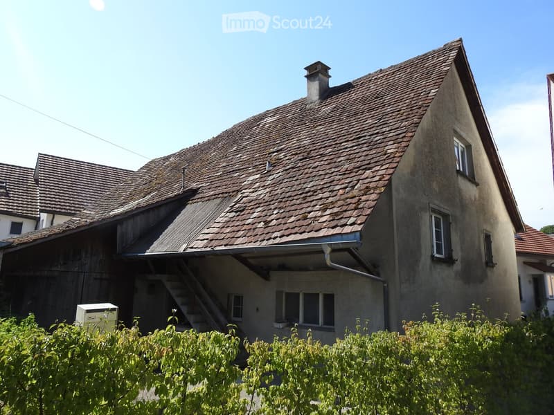 Two-story house with a tiled roof, chimney, and multiple windows. The house has a raised ground floor and is surrounded by greenery.