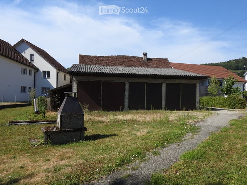 A grassy yard with a stone barbecue, a gravel path, and a large garage attached to a house with a red-tiled roof.