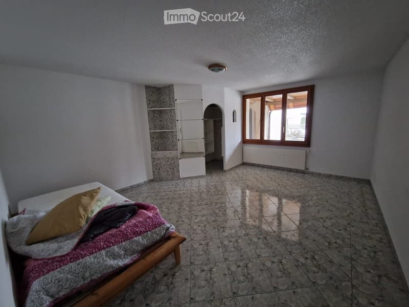 Empty bedroom with tiled floor, large window, and a bed with pillows and blankets.