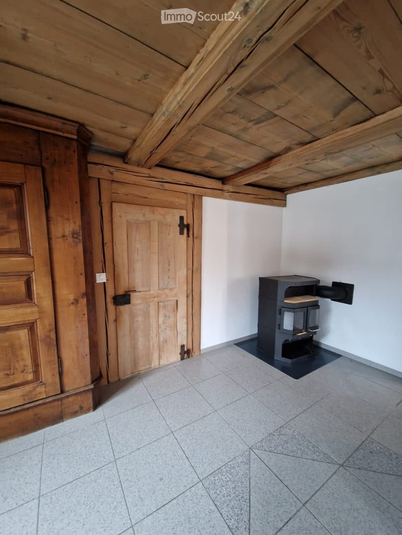 Empty room with exposed wooden beams on the ceiling, tiled floor, and a small wood-burning stove against the wall.
