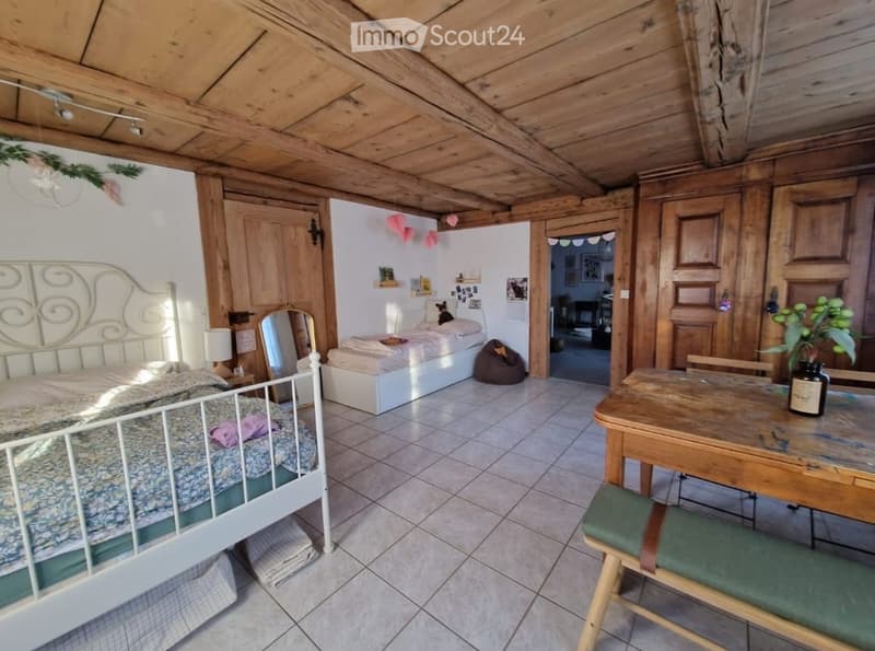Cozy bedroom with wooden ceiling and walls, featuring a double bed, a small table with a chair, a mirror, and a lamp. The room includes a second bed and a small seating area with a stool.