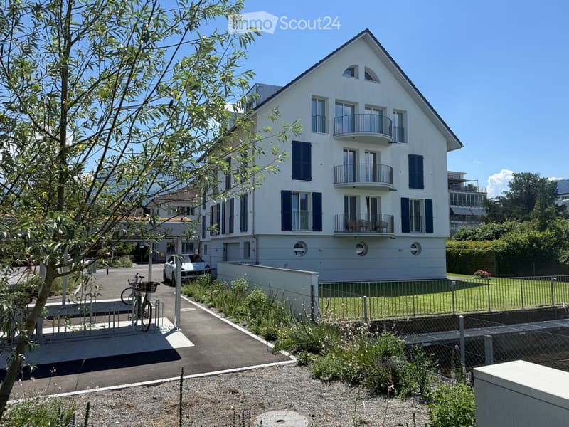 Modern two-story white building with multiple balconies and blue shutters, surrounded by greenery and a parking area.