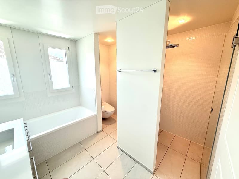 Modern bathroom with a bathtub, toilet, and separate shower area. Features tiled walls and floors, a window, and built-in storage cabinets.
