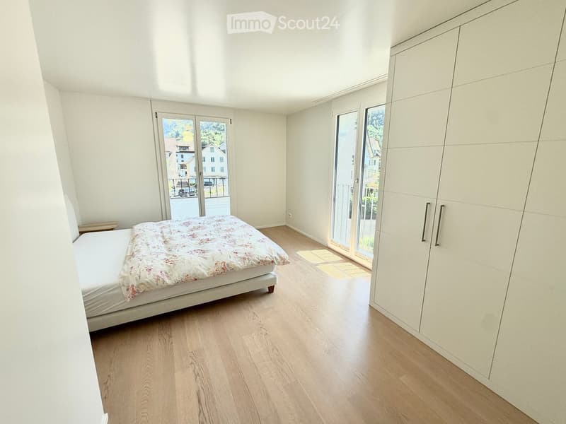 Bright bedroom with wooden flooring, a white bed with floral bedding, and a large built-in wardrobe. Two sets of glass doors lead to a balcony or outdoor area.