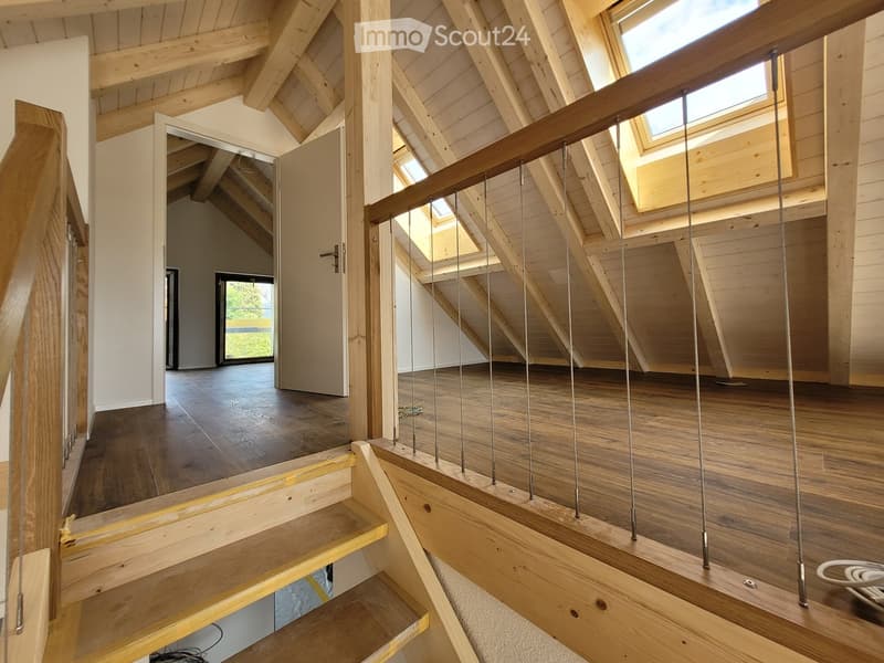 Spacious, open hallway with wooden floors, exposed wooden beams, and skylights, leading to a room with large windows and a dark wooden floor.