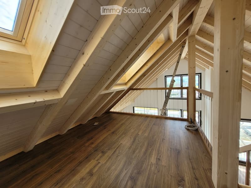 An empty attic space with exposed wooden beams and a wooden floor, featuring large windows and a modern metal railing along the edge.