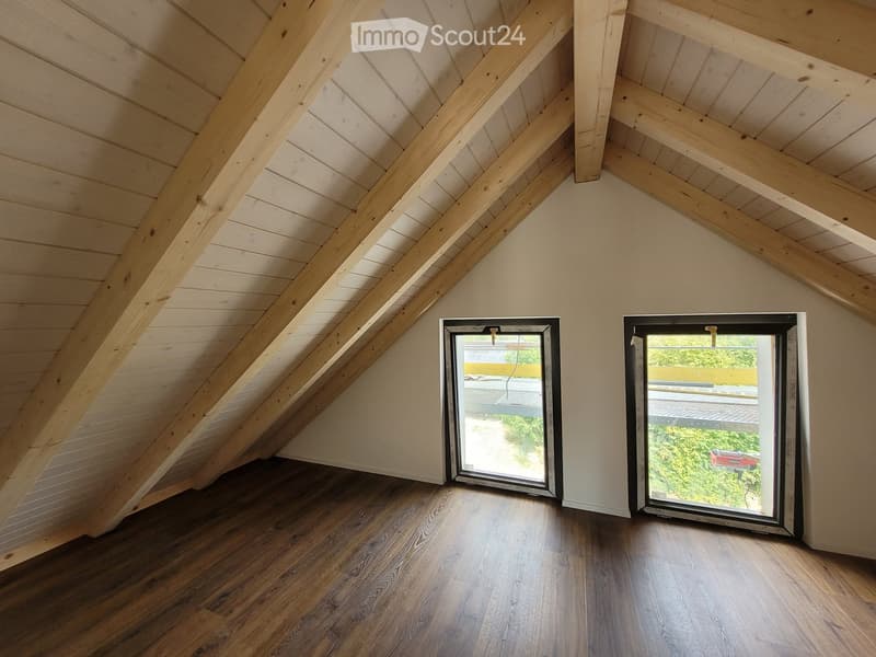 An empty attic space with wooden beams and flooring, featuring two large windows offering a view of the outdoors.