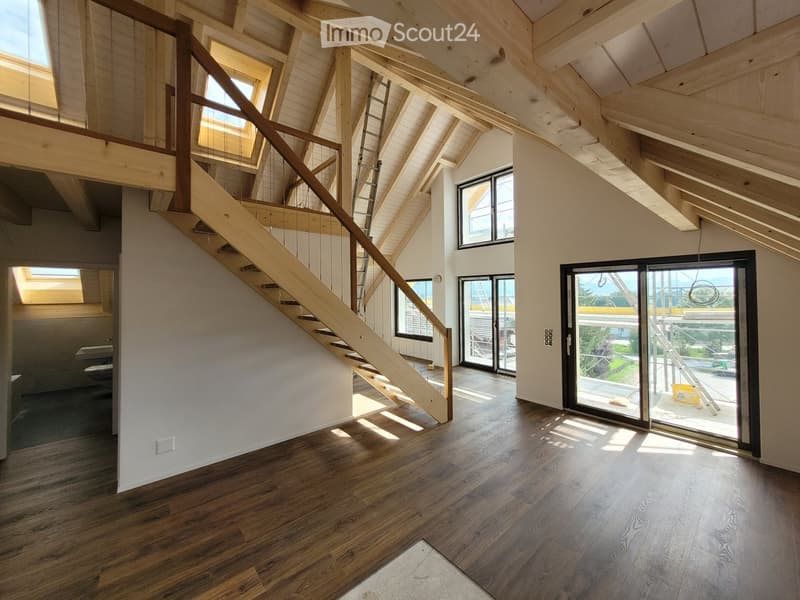 Open, airy space with wooden floors, exposed wooden beams, large windows, and a staircase leading to an upper level. No furniture or furnishings are present.