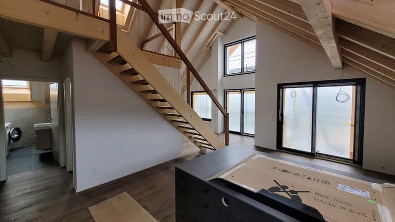 Empty room with wooden flooring, exposed wooden beams, and a staircase leading to an upper level. Large windows and sliding glass doors provide natural light.