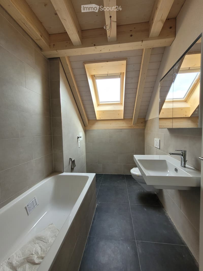 Modern bathroom with a bathtub, sink, and toilet, featuring grey tiles and wooden beams on the ceiling. Two skylights provide natural light.