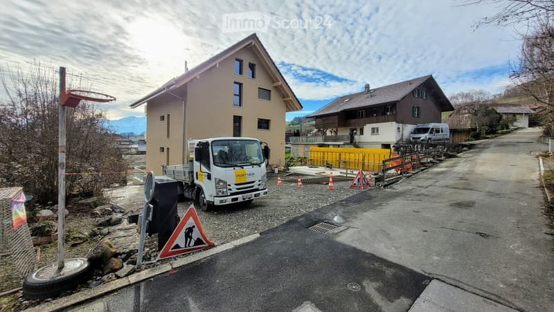 Two-story residential building with beige and brown exterior, located in a rural area with mountains in the background. A truck is parked nearby, and construction barriers and traffic cones are visible on the road.