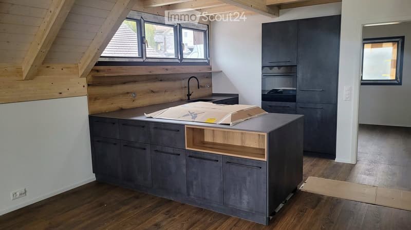 Modern kitchen with grey cabinets, wooden accents, and a central island. Exposed wooden beams on the ceiling and large windows provide natural light.