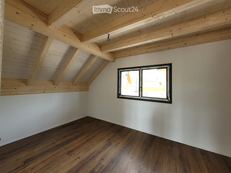 Empty room with wooden floor, white walls, and exposed wooden ceiling beams. A single window with a black frame is visible on one wall.