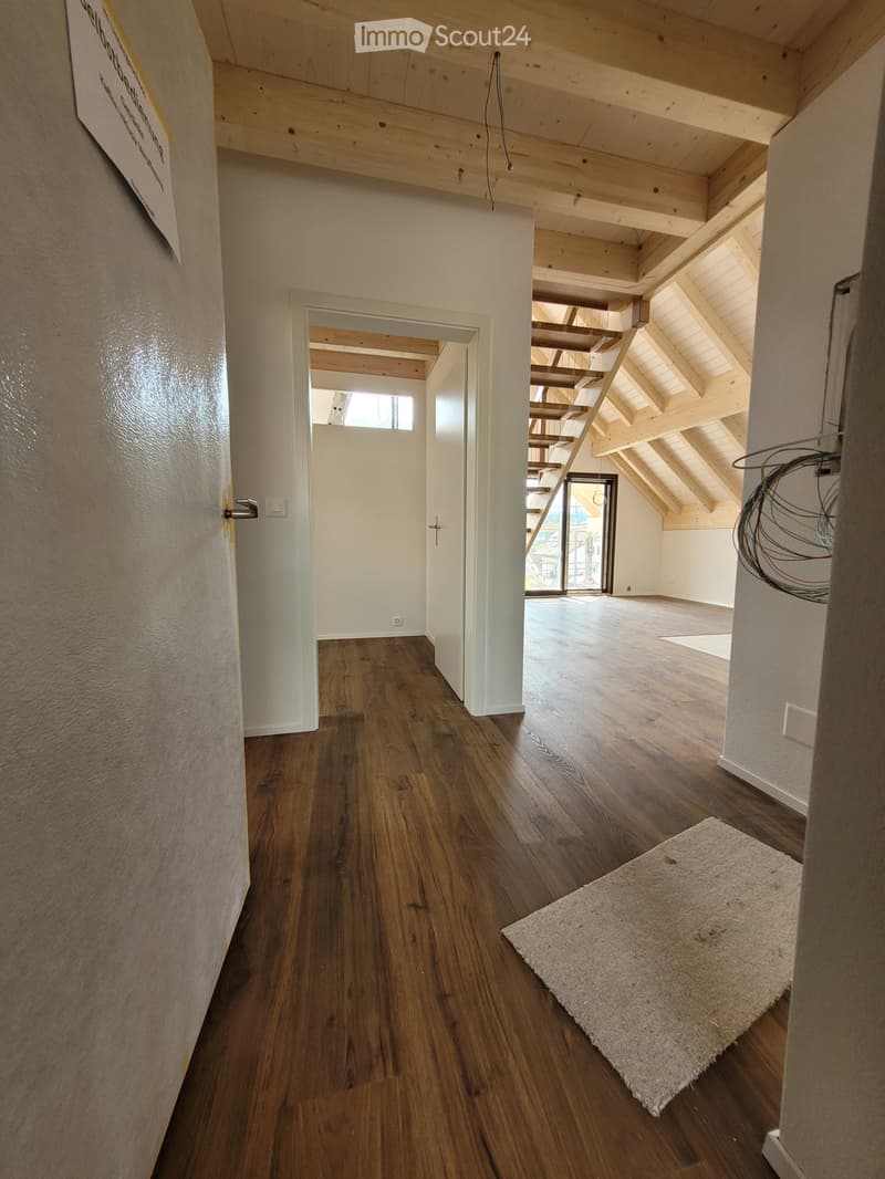 Empty hallway with wooden flooring, white walls, and exposed wooden beams in the ceiling. A small rug is placed near the doorway leading to another room.