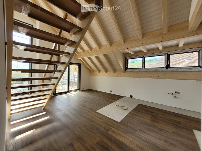 Empty room with wooden flooring, exposed wooden beams, and a staircase leading upwards. Large windows provide natural light, and the space is unfurnished.