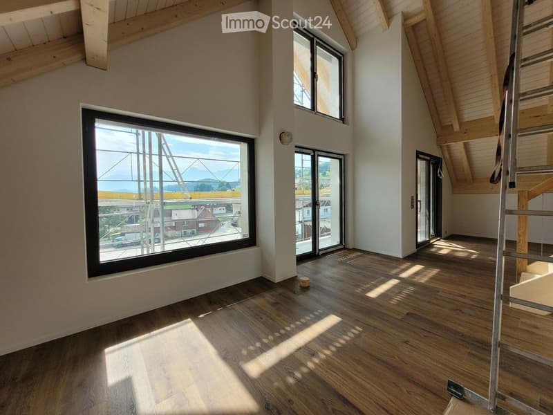 Empty room with wooden flooring, large windows, and exposed wooden beams on the ceiling. Natural light streams in, creating shadows on the floor.