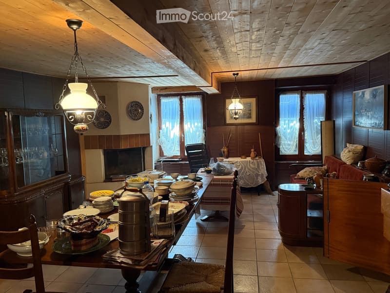 A cozy, rustic living and dining area with wooden beams, a fireplace, a large dining table set with dishes and utensils, a sofa, and a chandelier.