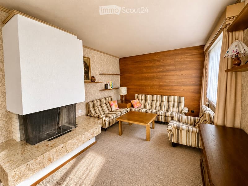 Cozy living room with a fireplace, patterned sofas, coffee table, armchair, and wooden shelves.