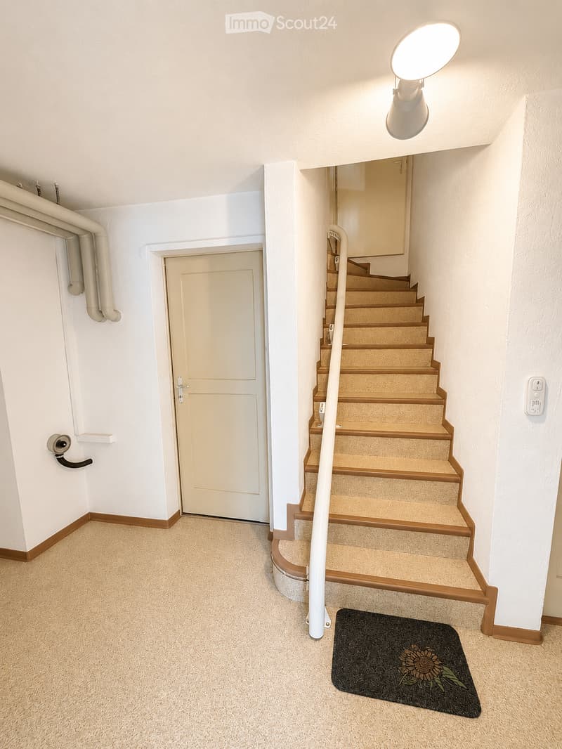 Carpeted hallway with a staircase leading upwards, a door on the left, and a small black doormat with a sunflower design near the stairs.