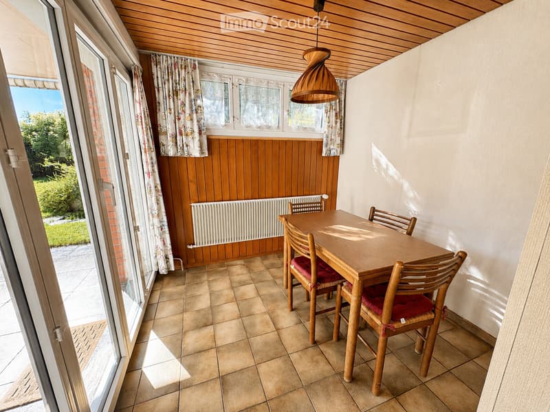 Brightly lit kitchen with wooden ceiling, tiled floor, and large windows. Includes a dining table with four chairs, radiator, and a hanging wooden pendant light.