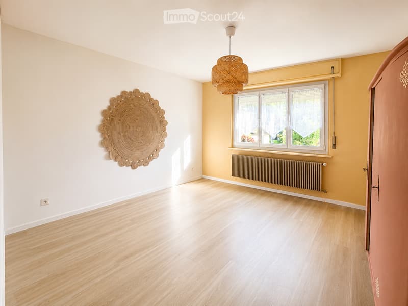 Empty room with light wooden flooring, white and yellow walls, a large window with white curtains, a radiator, and a decorative wall hanging. No furniture or equipment is present.
