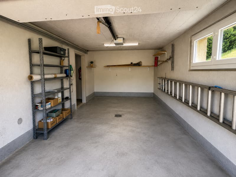 Empty garage with concrete floor, metal shelving unit, and a small window showing greenery outside.