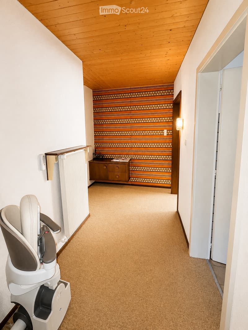 Carpeted hallway with wooden ceiling, white walls, and a small cabinet with drawers at the end. A baby chair is placed on the left side.