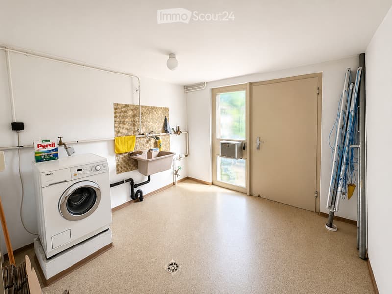 A clean, modern bathroom with a washing machine, a sink, a towel, and a detergent box. The space features white walls, a tiled backsplash, and a window with a view of greenery.
